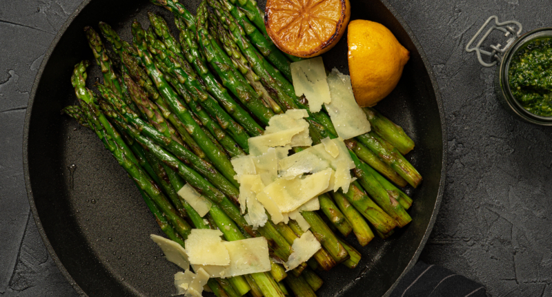 Fried green asparagus with lemon and parmesan in a pan. Black marble table with olive oil and pesto. Top view.